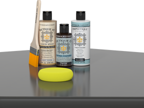 Retique It® Light Wood Wood’n Primer with Charcoal Gel Stain Alternative, gloss topcoat, brush, and yellow sponge on a reflective tabletop.