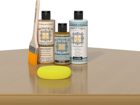 Retique It® Light Wood Wood’n Primer with Old Oak Gel Stain Alternative, gloss topcoat, brush, and yellow sponge on a tabletop.