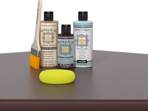 Retique It® Light Wood Wood’n Primer with Red Mahogany Gel Stain Alternative, satin topcoat, brush, and yellow sponge on a tabletop.