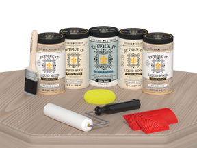 Retique It® Sun Bleached finishing system displayed on a light wood surface, featuring Liquid Wood Wood’n Primer in Bleached Wood, Liquid Wood Wood’n Stain in Pickled Oak, White Wash Gel Stain Alternative, paintbrush, roller, yellow sponge, and red graining tools for creating a weathered sun-bleached wood finish.