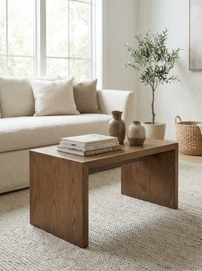 A modern wooden coffee table with a warm medium-brown finish, styled in a bright, neutral living room next to a cream sofa and a potted plant.