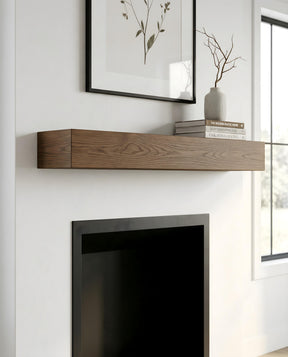 A floating wood mantel shelf mounted on a white wall above a black fireplace, styled with a framed botanical print, books, and a small vase with branches.