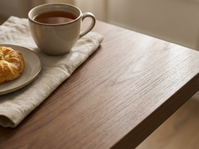 A warm, close-up lifestyle shot of a wooden table with a light beige ceramic cup of tea on a matching napkin, alongside a small plate with a croissant.