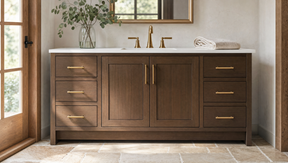 A dark brown wood bathroom vanity with a white countertop, gold faucets, and brass-toned drawer pulls, shown in a styled bathroom setting with a mirror, window, and stone tile floor.