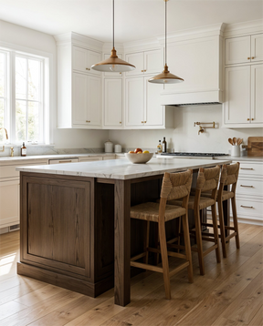 A bright, modern kitchen featuring a dark wood island with a white marble countertop and four woven-back bar stools. White shaker-style cabinets, brass pendant lights, and light wood flooring complete the scene.