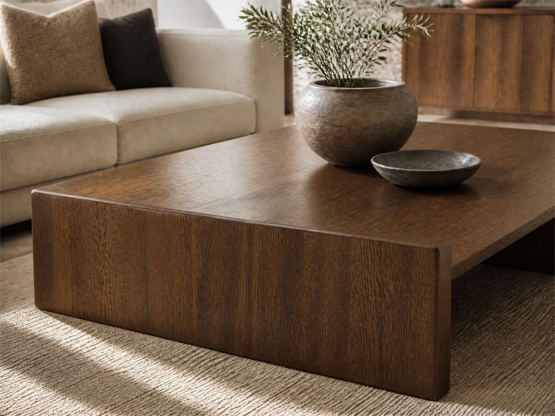 A modern low coffee table made of dark brown wood with a thick blocky base, styled in a living room setting with a beige sofa, a ceramic planter, and a small bowl on top.