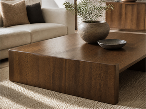A modern low coffee table made of dark brown wood with a thick blocky base, styled in a living room setting with a beige sofa, a ceramic planter, and a small bowl on top.