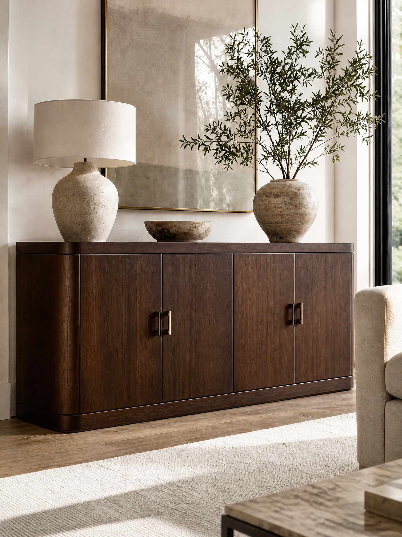 A dark brown wooden sideboard cabinet in a modern living room setting, styled with a textured ceramic table lamp, a small stone bowl, and a large vase with olive branches.