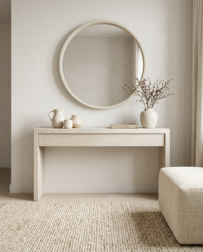 A minimalist interior scene featuring a round wall mirror with a light wood frame hanging above a matching light wood console table. The table is styled with ceramic vases and a vase of dried branches, set on a woven rug next to a textured beige ottoman.